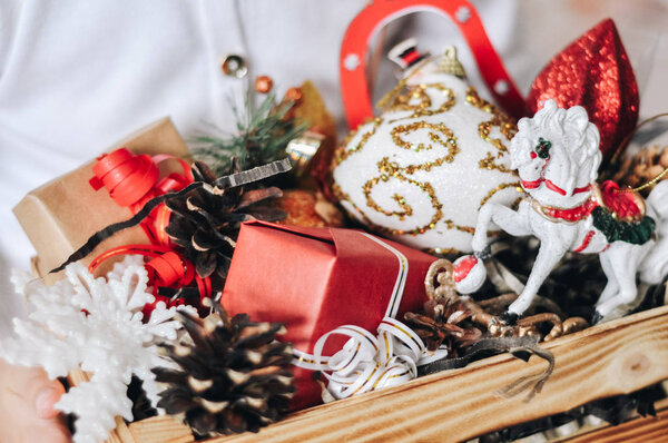 Деревянная коробка с рождественскими красочными различными игрушками и декором в руках маленькой девочки. Лошадь, звезды, конусы, снежинка, снеговик, мяч, подарки. Рождество, Новый год и украшение. Закрыть
