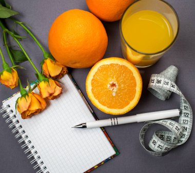 Not defteri ve kalem gri arka plan üzerinde. Portakal, turuncu gül, santimetre, bir bardak portakal suyu. Üstten Görünüm. Bir etiket için bir yer