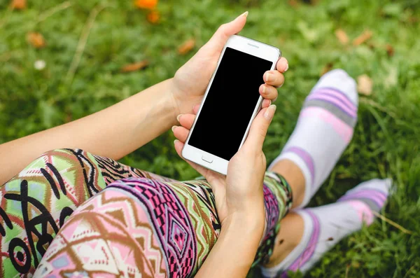 Fechar o telefone celular branco na moda, as mãos da mulher na moda no fundo da grama verde com flores durante uma pausa para exercícios — Fotografia de Stock