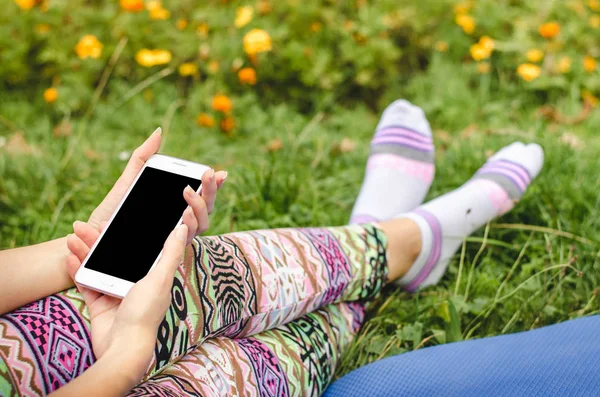 Fechar o telefone celular branco na moda, as mãos da mulher na moda no fundo da grama verde com flores durante uma pausa para exercícios — Fotografia de Stock