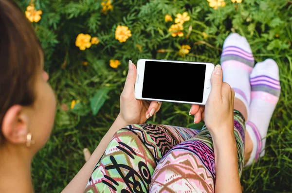 Fechar o telefone celular branco na moda, as mãos da mulher na moda no fundo da grama verde com flores durante uma pausa para exercícios — Fotografia de Stock