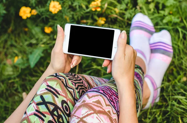 Fechar o telefone celular branco na moda, as mãos da mulher na moda no fundo da grama verde com flores durante uma pausa para exercícios — Fotografia de Stock