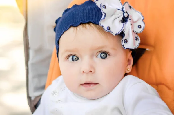 Портрет милой маленькой улыбающейся девочки с голубым цветочком-луком на голове на оранжевом фоне. Крупным планом ребенок смотрит в камеру с большими голубыми глазами . — стоковое фото