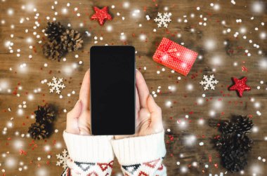 Kadın sıcak örme kazak Noel kar taneleri ve kar ile ahşap arka plan üzerinde akıllı cep telefonu ile oled göstermek tutan eller. Mutlu yeni yıl ve Xmas düz kompozisyon üstten görünüm yatıyordu.