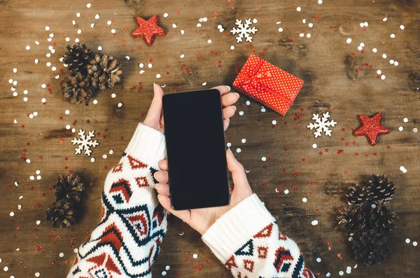 Female hands in a warm knitted sweater holding smart mobile phone with oled display on wooden background with Christmas gifts snowflakes and snow. Happy New Year and Xmas Flat lay composition top view.