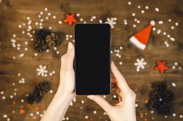 Female hands in a warm knitted sweater holding smart mobile phone with oled display on wooden background with Christmas gifts snowflakes and snow. Happy New Year and Xmas Flat lay composition top view.