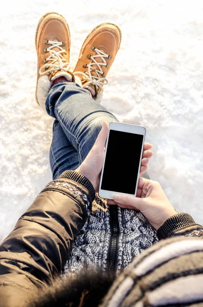 Hipster woman sitting in snow using white mobile smart phone, oled display, on wild nature, winter vacation, hiking, traveling, backpacker, warm clothes in Christmas time on a cold winter day