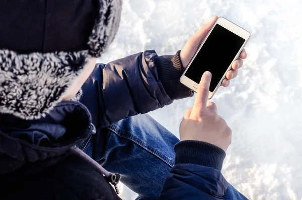 Hipster business man sitting in snow using white mobile smart phone, oled display, on wild nature, winter vacation, hiking, traveling, backpacker, warm clothes in Christmas time on a cold winter day
