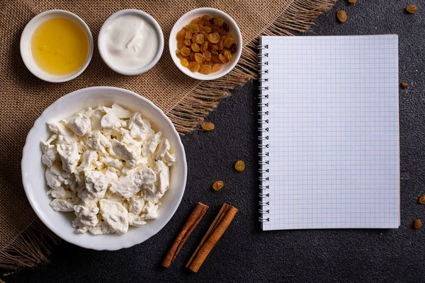 Beyaz tabakta süzme peynir, ayrı ayrı bal, kuru üzüm, ekşi krema, siyah arka planda beyaz bir defterin yanında. Tarifler için fotoğrafçılık.