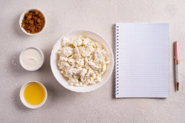 Beyaz tabakta süzme peynir, ayrı ayrı bal, kuru üzüm, ekşi krema, beyaz arka planda beyaz bir defterin yanında. Tarifler için fotoğraf. Üst görünüm.