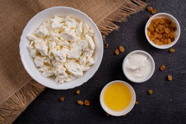 Beyaz tabakta süzme peynir, ayrı ayrı bal, çuval bezinde kuru üzüm, koyu bir arka plan. Üst görünüm.