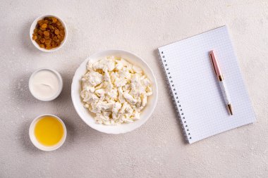 Beyaz tabakta süzme peynir, ayrı ayrı bal, kuru üzüm, ekşi krema beyaz arka planda beyaz bir defterin yanında. Tarifler için fotoğraf. Yukarıdan görüntüle.