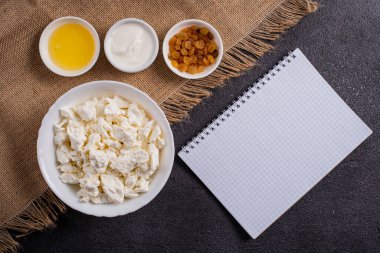 Beyaz tabakta süzme peynir, ayrı ayrı bal, kuru üzüm, ekşi krema, siyah arka planda beyaz bir defterin yanında. Tarifler için fotoğrafçılık.