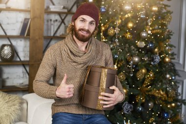 O sevinir ve Noel dekor Noel ağacına kış giyim moda adam arka plan ile rahat daire büyük bir başparmak gösterir onun hediye adam memnun