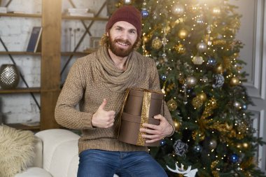 O sevinir ve Noel dekor Noel ağacına kış giyim moda adam arka plan ile rahat daire büyük bir başparmak gösterir onun hediye adam memnun