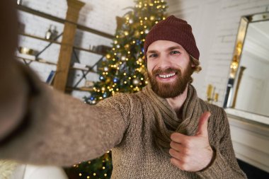 Yakışıklı genç adam Noel selfie ve gülümsüyor ağacı duruyor. Bir rahat yeni yıl daire moda adam. Genç adam fantezi kazaklı onun syrprise sunmak için hazır
