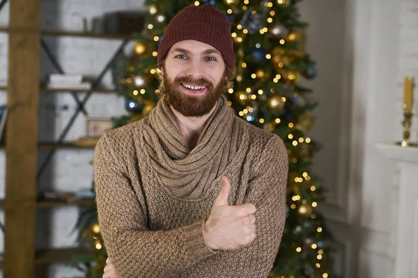 Fashionable male model thumbs up gesture. Christmas tree on background. Happy new year or merry xmas concept. Awesome emotions on party.