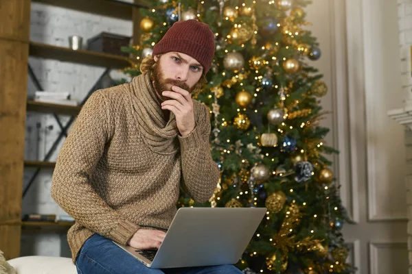 Hombre de Navidad en el portátil haciendo compras por Internet. Joven modelo masculino entusiasmado con la compra de regalos en línea en su computadora portátil. Navidad o Año Nuevo. Pensando en el concepto de regalo apartamento decorado . — Foto de Stock