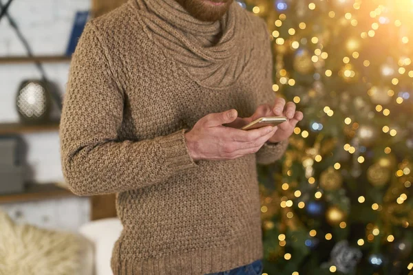 Hombre El uso de teléfono inteligente en la habitación decorada para Navidad. Buen bokeh. Compras en línea tema de año nuevo. Modelo irreconocible compra de regalo o mensajes de texto. Sms felicitaciones — Foto de Stock