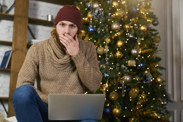 A fiatalember felejtett-hoz vesz egy ajándék karácsony vagy újév. A srác egy laptop karácsonyfa háttér. Hangulatos lakás. Fél felkészülés. Online vásárlás Stock Fotó