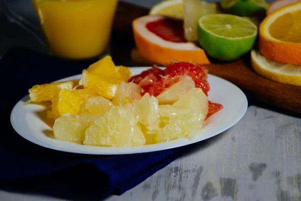 Varios cítricos cortados en rodajas naranja, limón, lima, pomelo, pomelo y un vaso de jugo de naranja. Extendido sobre una tabla de madera y un plato blanco vintage sobre el fondo de la textura de madera natural . — Foto de Stock