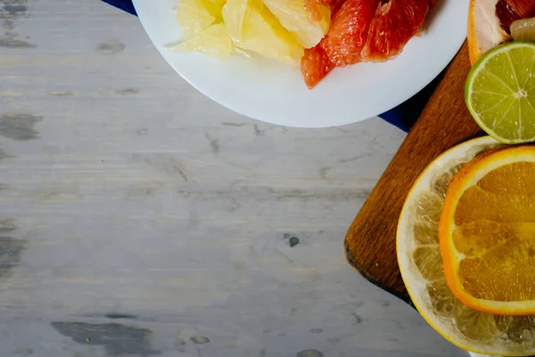 Varios cítricos cortados en rodajas naranja, limón, lima, pomelo, pomelo y un vaso de jugo de naranja. Extendido sobre una tabla de madera y un plato blanco vintage sobre el fondo de la textura de madera natural . — Foto de Stock