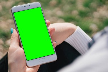 Kız ellerinde sahte yeşil ekran, boş ekran yukarı ile smartphone ile park açık alan, içerik entegrasyonu için ekran üzerinde oturuyor. Gadget'ı bulanık zemin üzerinde Önden Görünüm holding eller