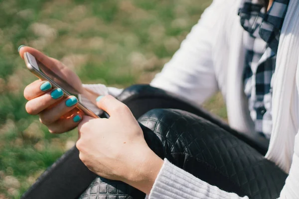 Hermosa joven europea se sienta en la hierba en el parque y utiliza un teléfono inteligente, conceptos de uso de aparatos en un entorno natural en el aire libre — Foto de Stock