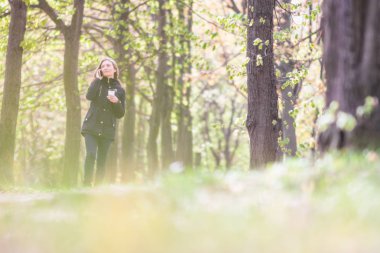 Bir kız sabah parkta elinde akıllı telefon ve kahveyle yürüyor.
