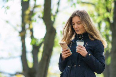 Bir kız sabah parkta elinde akıllı telefon ve kahveyle yürüyor.