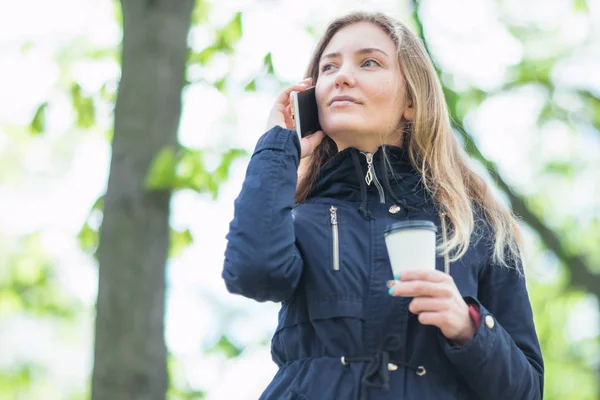 Hablando por teléfono, una chica está caminando por la mañana en un parque con un teléfono inteligente y café — Foto de Stock