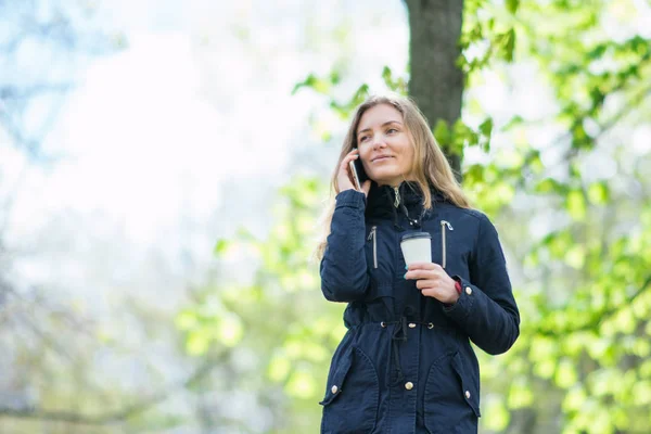 Hablando por teléfono, una chica está caminando por la mañana en un parque con un teléfono inteligente y café — Foto de Stock