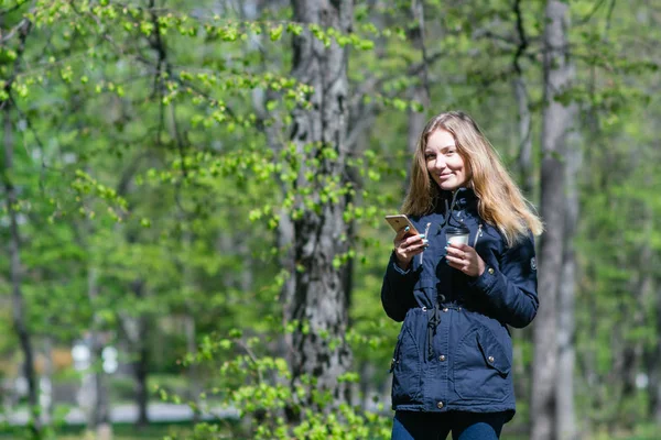 Una chica está caminando por la mañana en un parque con un teléfono inteligente y café — Foto de Stock