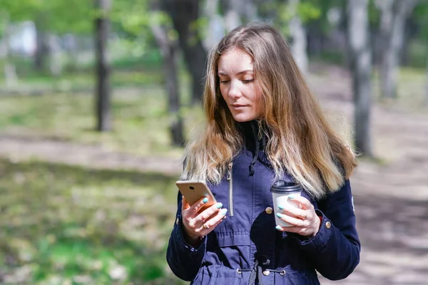 Una chica está caminando por la mañana en un parque con un teléfono inteligente y café — Foto de Stock