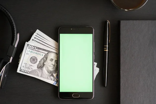 Smartphone with chromakey for keying and mockup on a black textured wooden table with One hundred dollar\'s banknotes of the USA. With pen, coffee, book and headphones