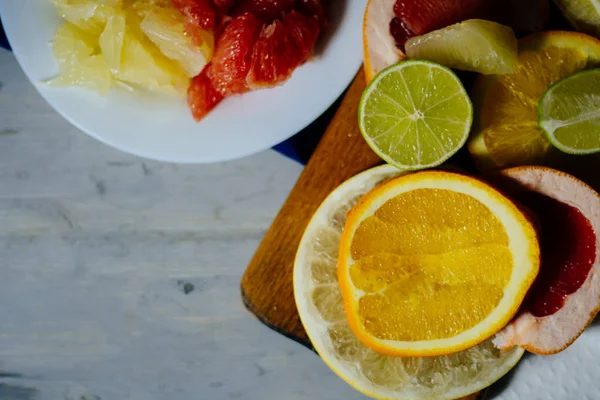 Varios cítricos cortados en rodajas naranja, limón, lima, pomelo, pomelo y un vaso de jugo de naranja. Extienda hacia fuera en una tabla de madera y un plato blanco vintage en el fondo de natural — Foto de Stock