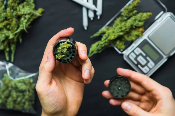 Un hombre usa un molinillo para moler marihuana con un fondo de flores de cannabis y escamas y una vista superior de las articulaciones —  Fotos de Stock