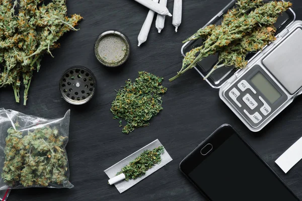 Cones de flores de maconha em escamas, moedor e junção de cannabis desfiada e um pacote de ervas daninhas e Smartphone em um fundo de madeira preta — Fotografia de Stock