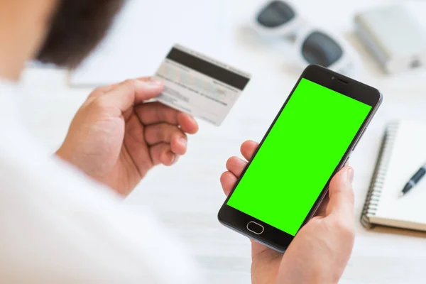 A black smartphone with green screen for chroma key compositing and a credit card in the hands of a man on a rural white background, concepts of Internet commerce and the use of online