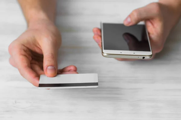 A black smartphone and a credit card in the hands of a man on a rural white background, concepts of Internet commerce and the use of online banking to pay for services and goods in Internet digital