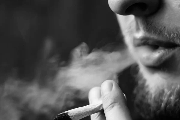 Un hombre fuma marihuana, un porro y un encendedor en sus manos. Humo sobre fondo negro. Conceptos de uso medicinal de la marihuana y legalización del cannabis.Blanco y negro — Foto de Stock