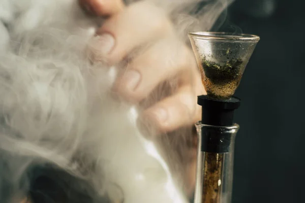 Raucht Cannabiskraut, eine Bong und ein Feuerzeug in seinen Händen. Rauch auf schwarzem Hintergrund. Konzepte des medizinischen Marihuana-Konsums und der Legalisierung von Cannabis. — Stockfoto