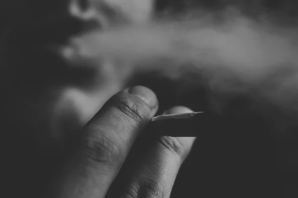 Un homme fume de l'herbe de cannabis, un joint et un briquet dans ses mains. Fumée sur fond noir. Concepts de la consommation de marijuana médicale et de la légalisation du cannabis.Noir et blanc — Photo