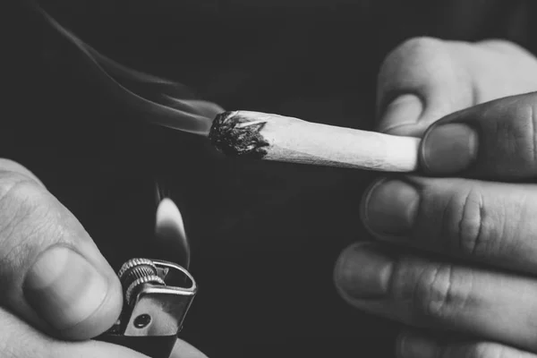 Um homem fuma erva de cannabis, um charro e um isqueiro nas mãos. Fumar em um fundo preto. Conceitos de uso medicinal de maconha e legalização do canabis.Preto e branco — Fotografia de Stock