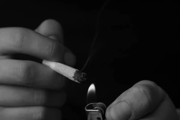 Más ligero y articulado en sus manos. Un hombre fuma marihuana, una. Humo sobre fondo negro. Conceptos de uso medicinal de la marihuana y legalización del cannabis.Blanco y negro — Foto de Stock