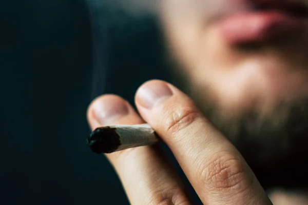 Hierba de cannabis, un porro en sus manos Un hombre fuma. Humo sobre fondo negro. Conceptos de uso médico de la marihuana y legalización del cannabis . — Foto de Stock