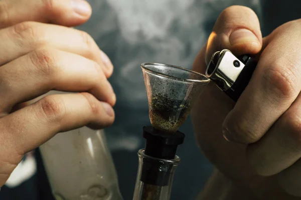 Bong e um isqueiro nas mãos. Um homem fuma erva de cannabis, um fumo num fundo preto. Conceitos de uso medicinal de maconha e legalização da cannabis . — Fotografia de Stock