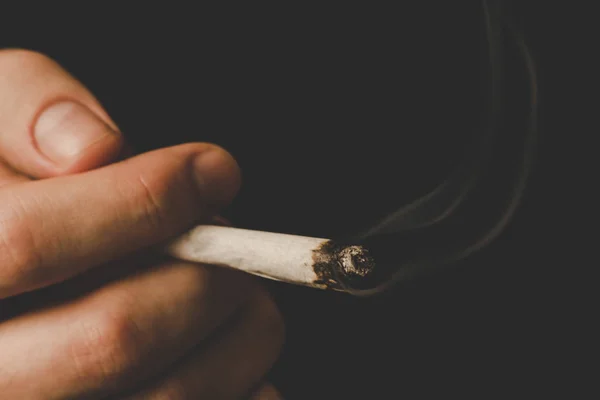 Hierba de cannabis, un porro en sus manos Un hombre fuma. Humo sobre fondo negro. Conceptos de uso médico de la marihuana y legalización del cannabis . — Foto de Stock