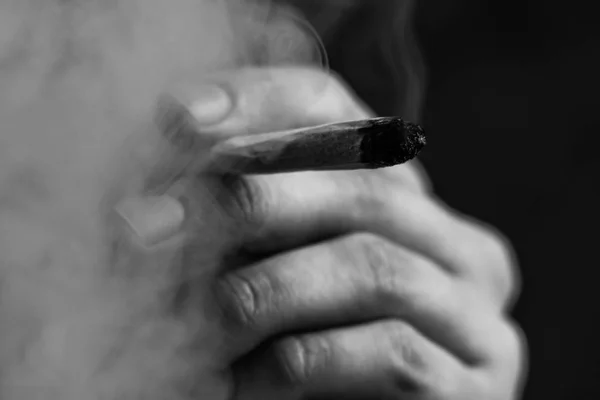 Smokes cannabis weed, a joint and a lighter in his hands. Smoke on a black background. Concepts of medical marijuana use and legalization of the cannabis.Black and white — Stock Photo, Image