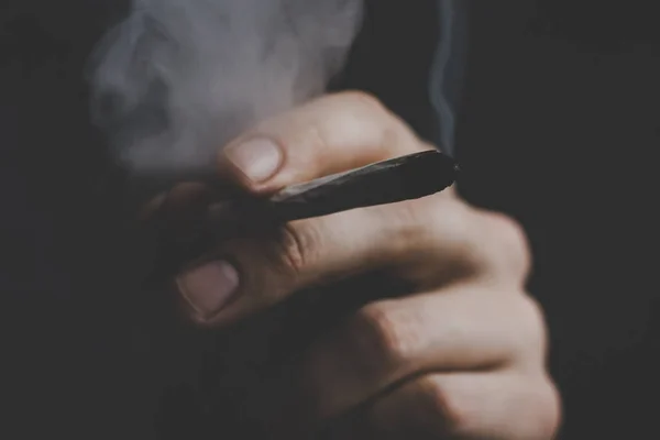 Спільний в руці людини. Чоловік курить бур'яни конопель. Дим на чорному тлі. Концепції медичного вживання марихуани та легалізації канабісу . — стокове фото